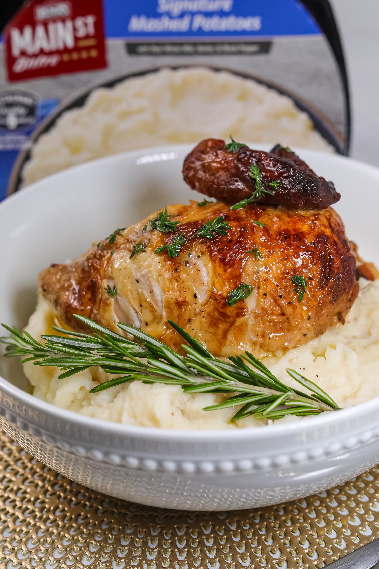 Roast chicken sitting in a bowl on top of Main St Bistro Signature Mashed Potatoes with rosemary sprig.